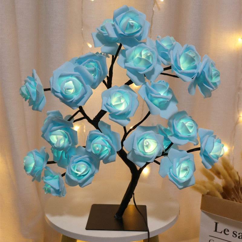 Árvore decorativa com flores azuis iluminadas em mesa redonda branca, fundo com cortina bege e luzes acolhedoras