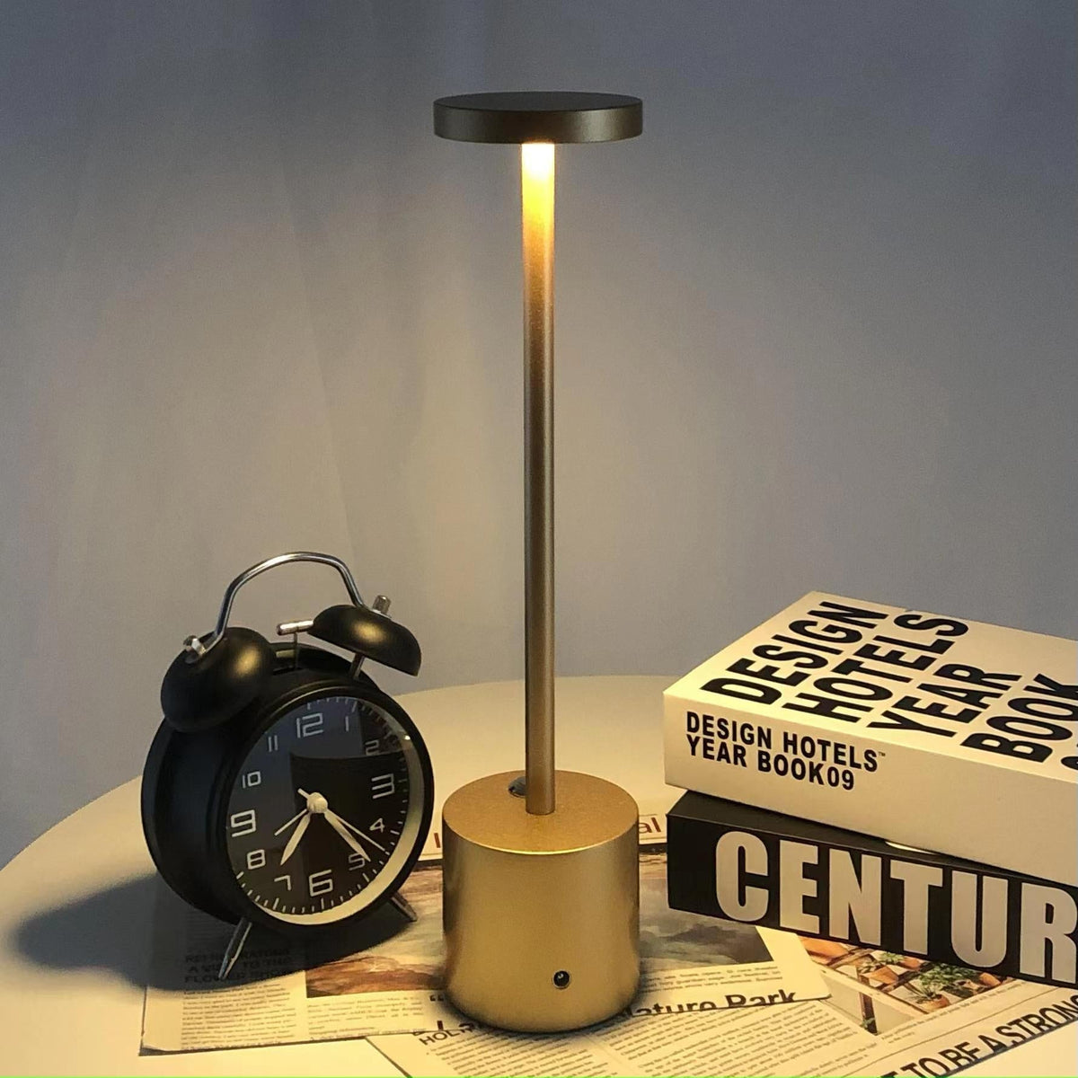 Relógio despertador preto, luminária dourada acesa em mesa branca com livros de design