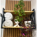 Prateleira de madeira com estrutura preta contendo toalhas enroladas e vasos com plantas decorativas